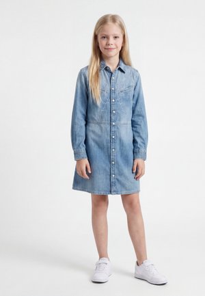 Ung pige med langt blondt hår iført en lyseblå denimkjole og hvide sneakers, stående mod en hvid baggrund.