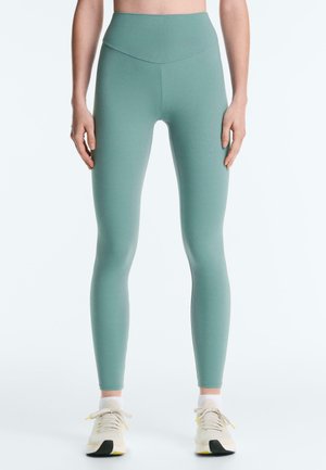 Femme portant des leggings taille haute vert mousse marine et des chaussures de sport blanches, debout devant un fond uni.