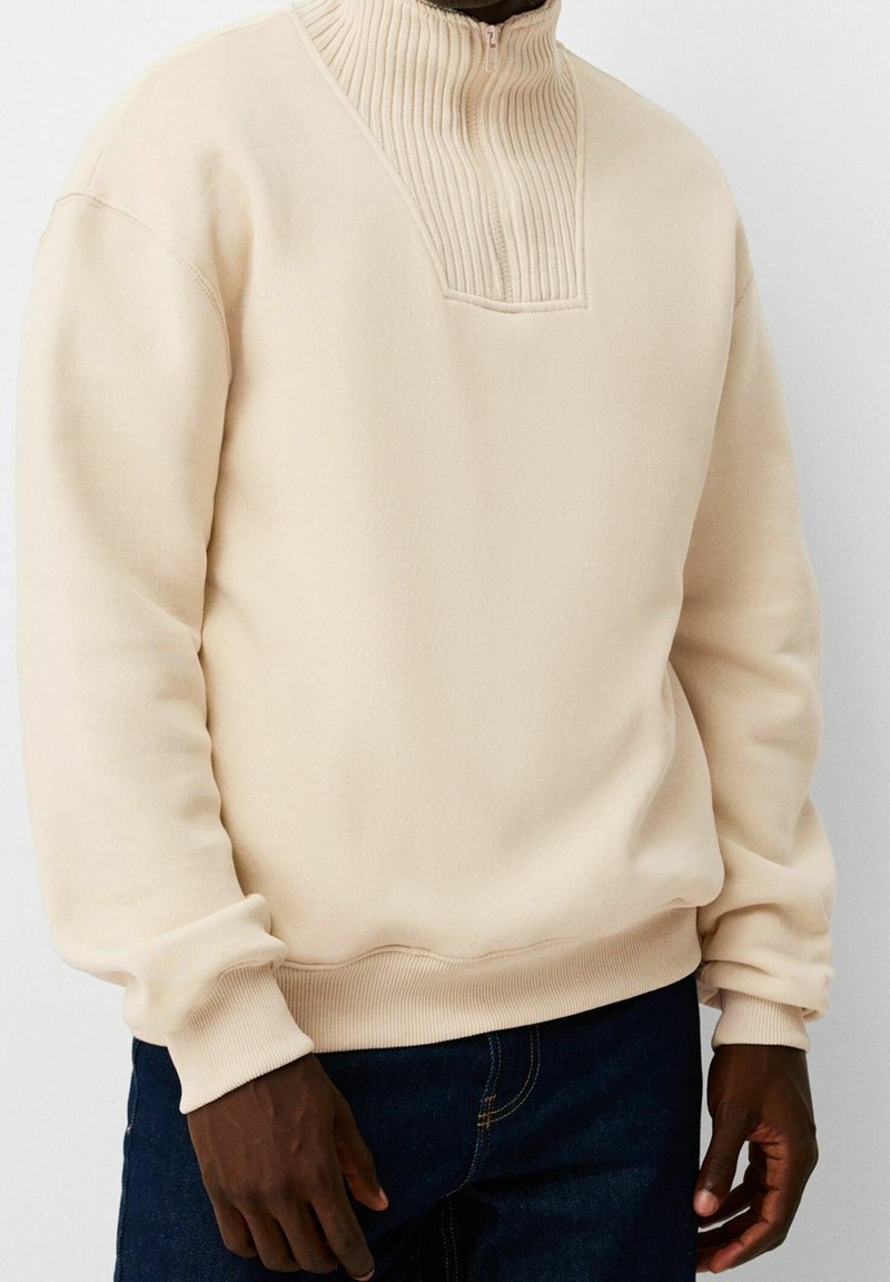 Pull beige avec col montant côtelé et détail en forme de V porté par une personne en jean bleu foncé, mains détendues sur les côtés.