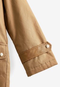 Manche d'une veste en tissu marron clair avec un poignet cousu et une sangle réglable à fermeture pression sur fond blanc.