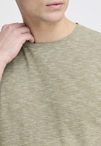 Olivengrøn t-shirt i et tekstureret, let meleret stof. Har rund hals og korte ærmer, vist på en person der holder i kraven.