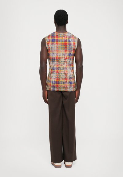 Modello maschile in top senza maniche a quadri multicolori, pantaloni larghi scuri e scarpe di colore chiaro, in piedi di spalle su sfondo neutro.