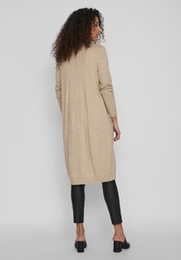 Long cardigan beige en tricot avec des manches longues et un ourlet côtelé, associé à un pantalon noir ajusté et des chaussures à talons épais. Texture lisse.