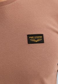 Tričko s krátkým rukávem, fitted střihu, vyrobené z bavlny v jemně růžové barvě. Obsahuje obdélníkovou černou nášivku s žlutým textem a detaily křídla.