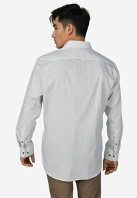 Ein Mann, der mit dem Rücken zur Kamera steht, trägt ein weißes Hemd mit langen Ärmeln, Knöpfen vorn und einem dezenten Punktmuster sowie braune Hosen vor einem schlichten Hintergrund.