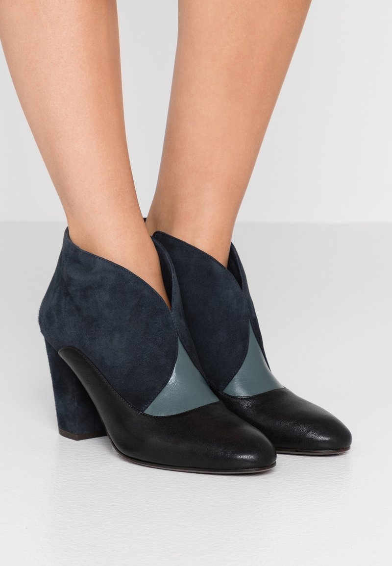 Bottines à talons hauts en cuir et suède noir, bleu foncé et gris portées pieds nus sur un fond neutre.
