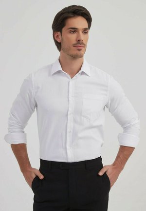 Chemise blanche à boutons avec une poche, manches longues retroussées au niveau des coudes, associée à un pantalon noir. Tissu lisse et design de col classique.