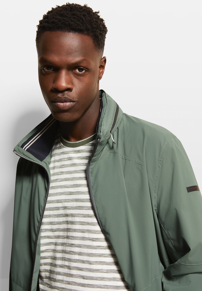 Veste légère verte avec un col haut, fermeture éclair et accents rayés. Portée par-dessus un t-shirt rayé blanc et gris.