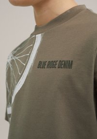 Alyvuogių žalių medvilnės marškinėlių, kurių priekyje yra balta grafika su ratu ir tamsiai žalia tekstas "BLUE RIDGE DENIM". Apvalus kaklas.