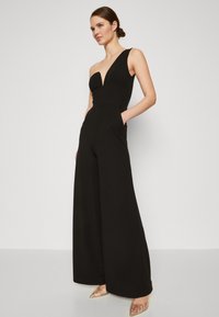 Svart jumpsuit med dyp V-hals, en skulderstropp, vid ben-design og sidelommer. Modellen har på seg spisse, transparente hæler.