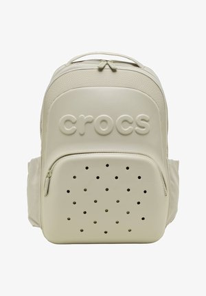 Beiger Crocs-Rucksack mit vorderer Reißverschlusstasche, die Belüftungslöcher und ein geprägtes Crocs-Logo auf einer glatt strukturierten Oberfläche aufweist.