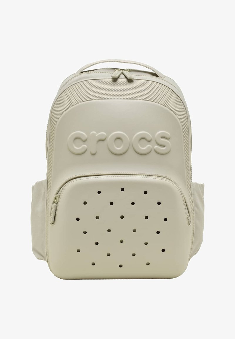 Beige Crocs-rugzak met voorvak met ritssluiting, voorzien van ventilatiegaatjes en een geperst Crocs-logo op een gladde, getextureerde oppervlak.