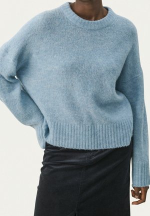 Person iført en lyseblå, blød strikket sweater med lange ærmer og et mørkt skørt, stående mod en ensfarvet lys baggrund.