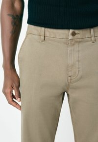 Close-up of a person wearing beige pants and a black ribbed shirt, showing a tattoo on the left forearm and hand resting by the side.