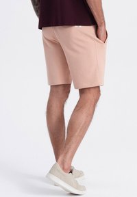 Shorts rose clair en tissu doux, avec des poches latérales et une coupe droite. Portés avec des baskets beiges, affichant un style décontracté.