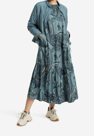 Vrouw draagt een teal midi-jurk met paisleyprint, een teal doorgestikte jas en stevige witte sneakers, staand met handen in de zakken van de jurk.