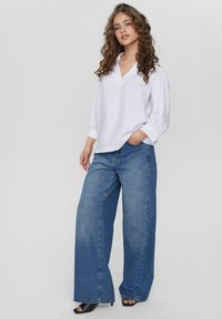Camicia bianca a maniche lunghe con scollatura a V, abbinata a jeans di denim blu a gamba larga. Sandali neri con tacco; capelli ricci e mani in tasca.
