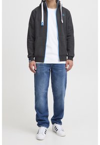 Grijze ritshoodie met blauwe accenten en witte koorden, gedragen met lichtblauwe jeans en witte sneakers met zwarte strepen.