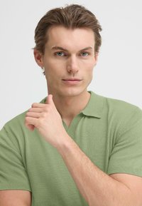 Polo à manches courtes vert clair avec un col classique et une texture en maille fine. La main du mannequin repose contre le menton. Fond neutre.