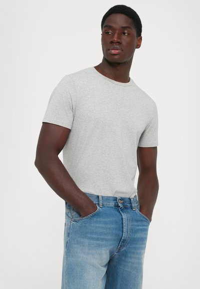 Uomo che indossa una maglietta a maniche corte grigio chiaro e jeans blu, in piedi con le mani in tasca, guardando di lato contro uno sfondo bianco semplice.