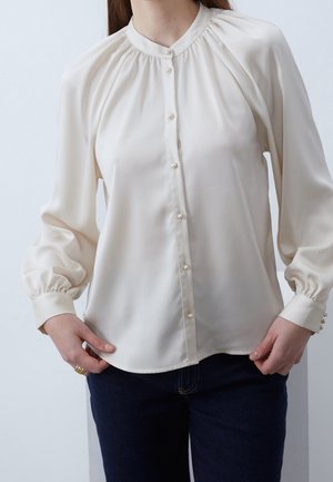 Nainen pukeutuneena kermaiseen pitkähihainen puseroon helmipainikkeilla ja tummansinisiin farkkuihin, kädet lepäävät lantiota vasten yksinkertaisen taustan edessä.