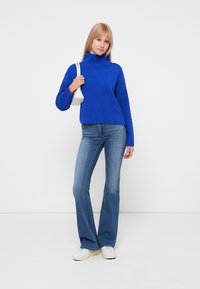 Un pull à col roulé tricoté bleu avec des détails texturés, associé à un jean flare bleu clair et des baskets blanches, tenant un petit sac à main blanc.