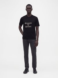 Άνδρας όρθιος, φοράει μαύρο T-shirt με το λογότυπο Emporio Armani, μαύρο τζιν και μαύρα αθλητικά παπούτσια πάνω σε απλό λευκό φόντο.