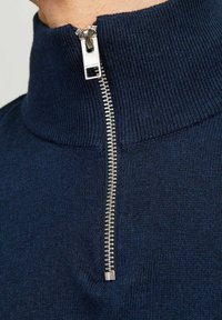 Granatowy sweter zapinany na zamek z wysokim kołnierzem i srebrnym zamkiem. Żebrowana struktura przy kołnierzu oraz gładkie ciało. Zbliżenie na dekolt.