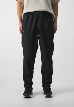 Mann trägt schwarze Under Armour Trainingshose mit Kordelzug in der Taille und schwarze Sportschuhe, steht vor einfachem Hintergrund.