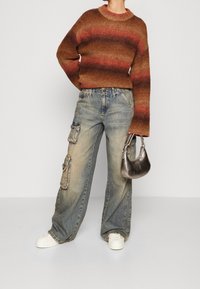 Suéter de punto a rayas marrón y rojo, vaqueros cargo azules holgados con bolsillos de parche, zapatillas blancas y un pequeño bolso de mano plateado.