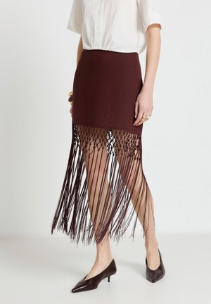 Femme portant une jupe marron taille haute avec de longues franges, associée à un chemisier blanc à manches courtes et des chaussures marron à petits talons.