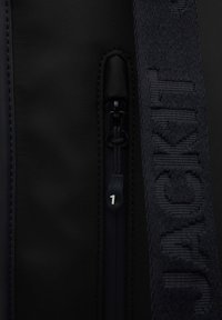 Borsa nera con una texture liscia, dotata di chiusura con zip e di una tracolla contrassegnata con il numero "1" in lettere rialzate e intrecciate lungo tutta la sua lunghezza.