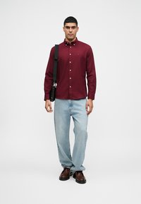 Burgundy-painu paita me logo, vaaleansiniset löysät farkut ja ruskeat saappaat. Henkilö kantaa mustaa laukkua olkapäällä.