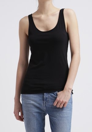 Frau trägt ein eng anliegendes schwarzes ärmelloses Tanktop und hellblaue Jeans, steht vor einem schlichten grauen Hintergrund.