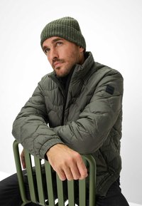 Olivengrøn puffjakke med struktureret overflade, høj krave og lynlås. Ribbede grønne hue. Siddende på en grøn metalstol.