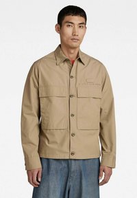 Giacca beige leggera con due grandi tasche sul petto, chiusura con bottoni e marchio discreto sul lato sinistro. Tessuto liscio, maniche lunghe.