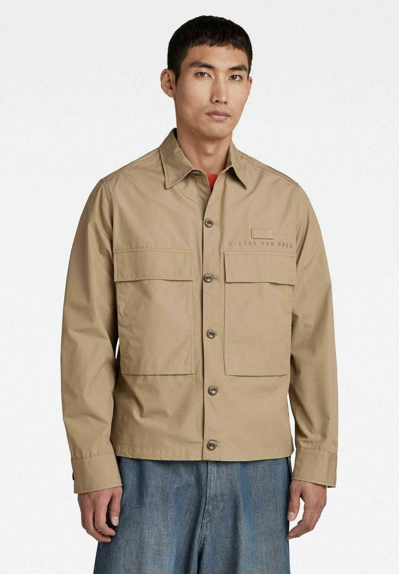 Giacca beige leggera con due grandi tasche sul petto, chiusura con bottoni e marchio discreto sul lato sinistro. Tessuto liscio, maniche lunghe.