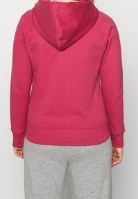 Persona che indossa un cappuccio rosso e pantaloni grigi, vista da dietro su uno sfondo bianco uniforme.