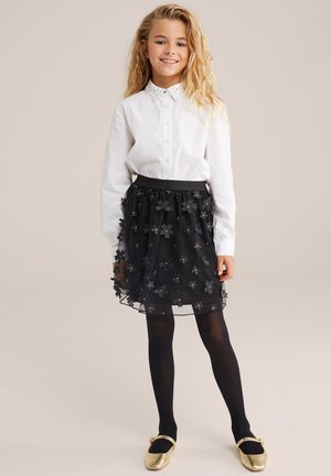 Cămașă albă cu guler brodat, purtată împreună cu o fustă din tulle negru decorată cu aplicații florale, colanți negri și pantofi aurii.