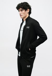 Μαύρο μπουφάν με φερμουάρ και ψηλό γιακά, συνδυασμένο με παντελόνι ίδιου υφάσματος. Διαθέτει λογότυπο στο στήθος και τσέπες. Το ύφασμα φαίνεται μαλακό και λείο.