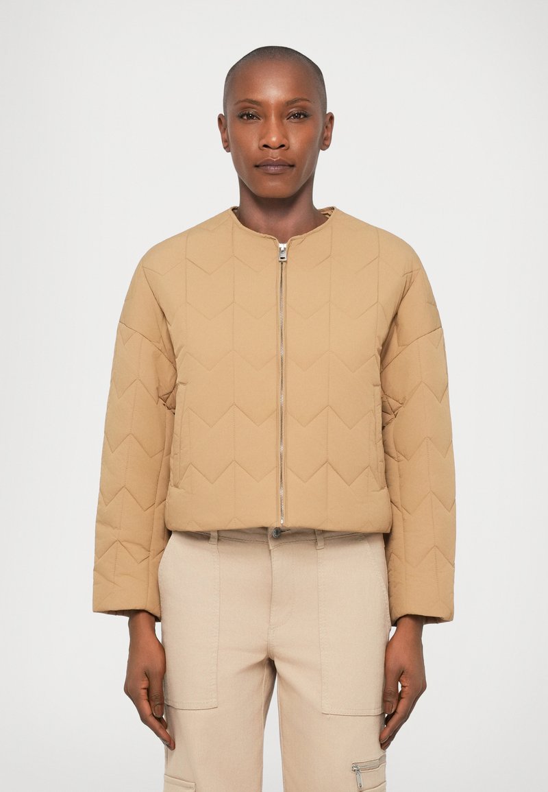 Giacca trapuntata beige con scollatura rotonda e zip anteriore. Presenta un motivo geometrico con un design corto. Tessuto morbido e vestibilità rilassata.