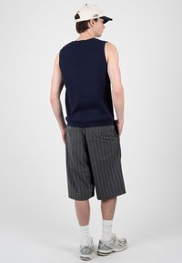 Homme portant un pull sans manches bleu marine, un short gris à fines rayures, des chaussettes blanches, des baskets et une casquette de baseball bicolore, debout les mains dans les poches.