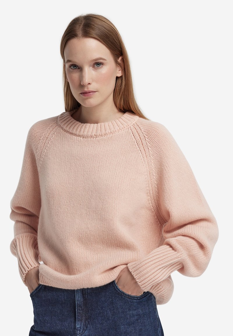 Rosa stickad tröja med rund halsringning, raglanärmar och ribbade muddarna, visas tillsammans med avslappnade jeans i denim. Mjuk textur och ledig passform.