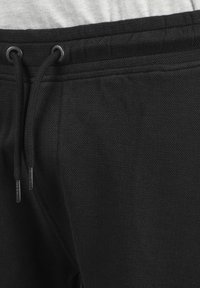 Pantalons de survêtement noirs en tissu texturé, dotés d'une taille avec cordon de serrage et d'œillets en métal. La vue en gros plan montre une apparence douce et tissée.