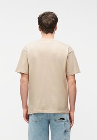 Άνδρας που φοράει μπεζ κοντομάνικη μπλούζα και ανοιχτόχρωμο μπλε τζιν, γυρισμένος πλάτη, μπροστά σε απλό λευκό φόντο.