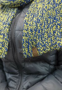 Scarf in maglia giallo e blu, caratterizzato da un motivo testurizzato. Accentuato con un'etichetta in pelle marrone. Indossato sopra un giubbotto nero imbottito.