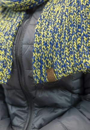 Scarf in maglia giallo e blu, caratterizzato da un motivo testurizzato. Accentuato con un'etichetta in pelle marrone. Indossato sopra un giubbotto nero imbottito.