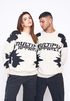 Sudaderas de color crema con patrones abstractos en negro y el texto "MULTIPLY APPAREL". Cuello redondo, mangas largas, corte relajado. Combinando con pantalones negros.