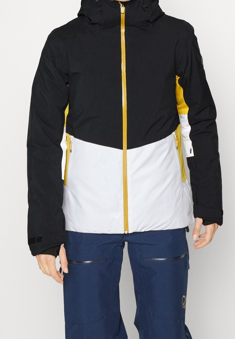 Veste colorée en blocs noir, jaune et blanc, avec capuche, fermeture éclair à l'avant et poches zippées ; fabriquée en matériau texturé et imperméable.