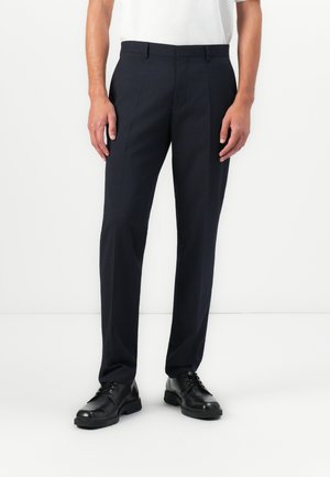 Man wearing tailored navy dress pants, black leather lace-up shoes, and a white tucked-in shirt on a plain white background.